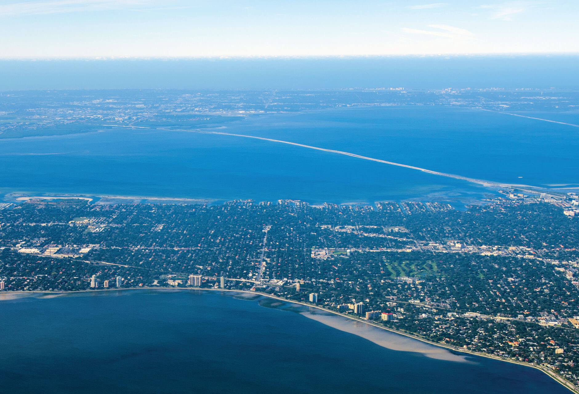 Aerial view across Tampa Bay showing the long Howard Frankland Bridge causeway linking Tampa in the foreground to St. Petersburg in the distance.