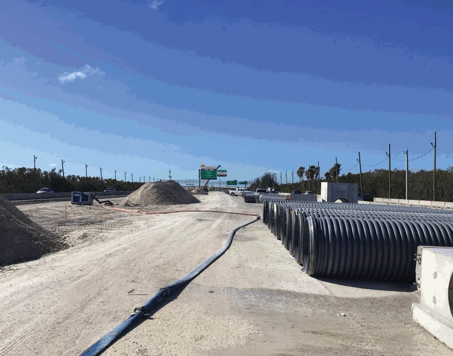 Rows of large black corrugated stormwater pipe and concrete drainage structures staged on a graded causeway work site beside highway traffic.