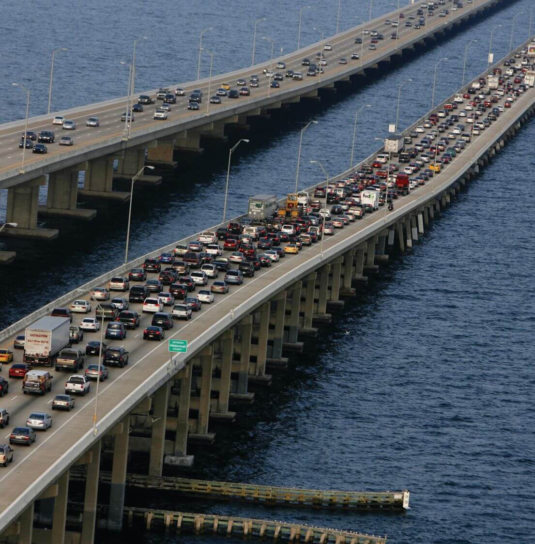 Aerial view of two parallel bridge spans over Tampa Bay, one packed with slow traffic beside an Entering Hillsborough County sign.