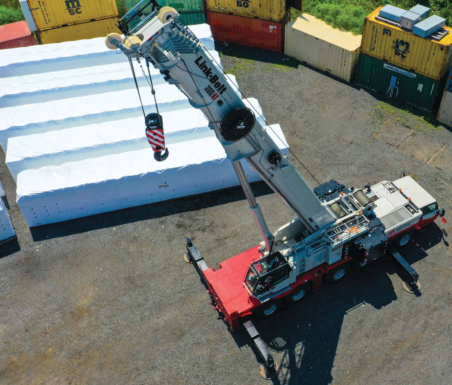 Overhead view of a red and white Link-Belt 300|AT all-terrain crane with outriggers set beside white-wrapped loads in a container yard.