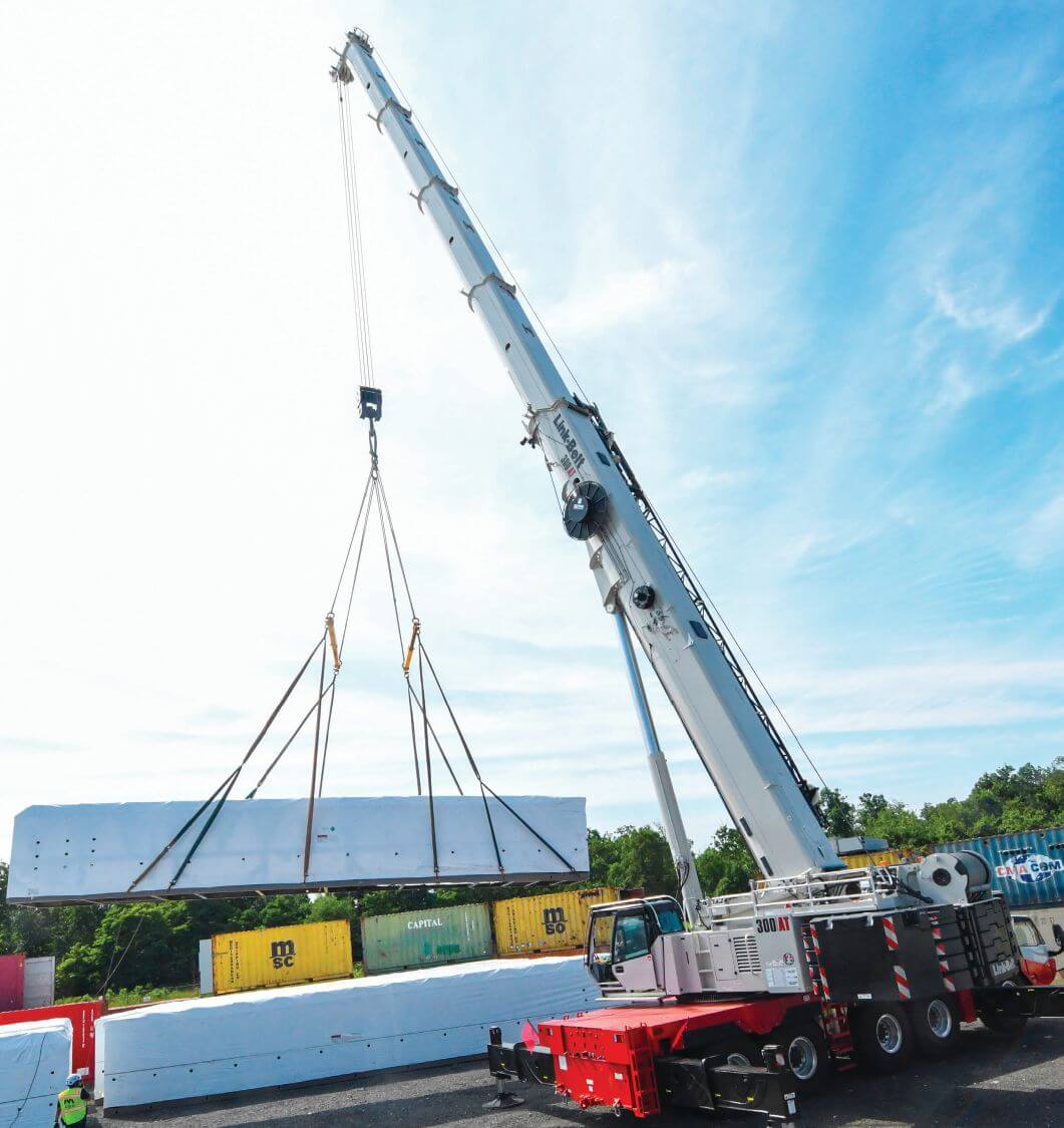 Link-Belt 300|AT crane with its boom extended lifting a long white-wrapped load on a multi-leg rigging in a yard lined with shipping containers.
