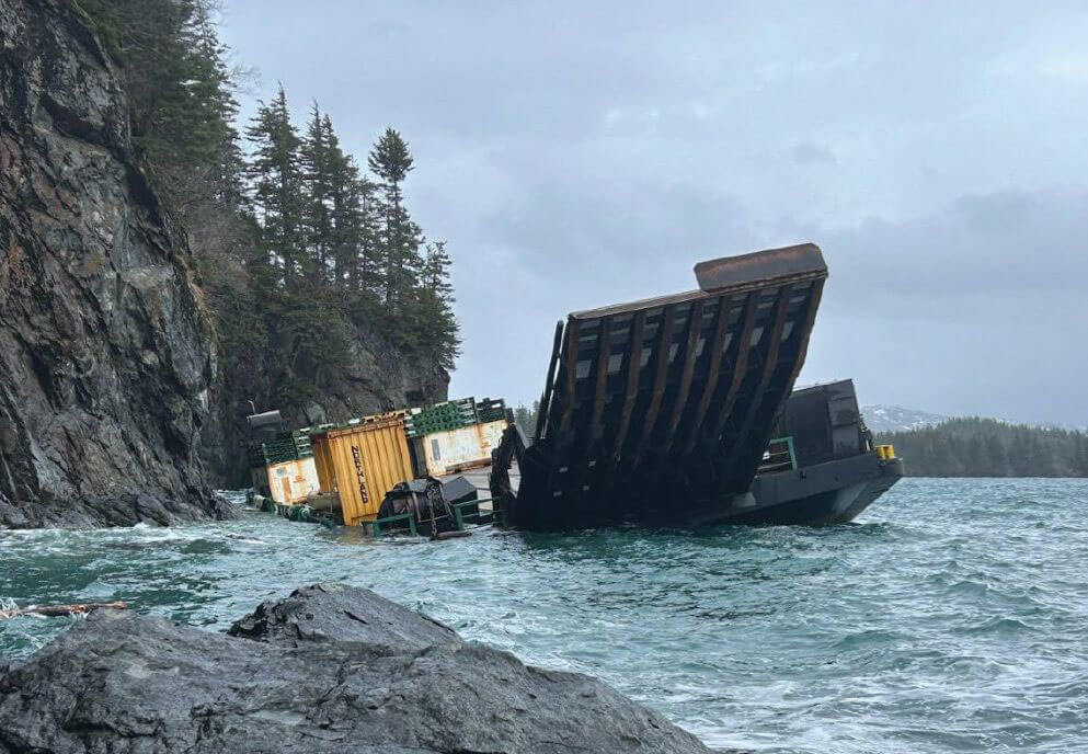 The barge Cordova Provider aground against a steep, tree-topped rock shoreline on Axel Lind Island, its containers and bow ramp in rough surf.