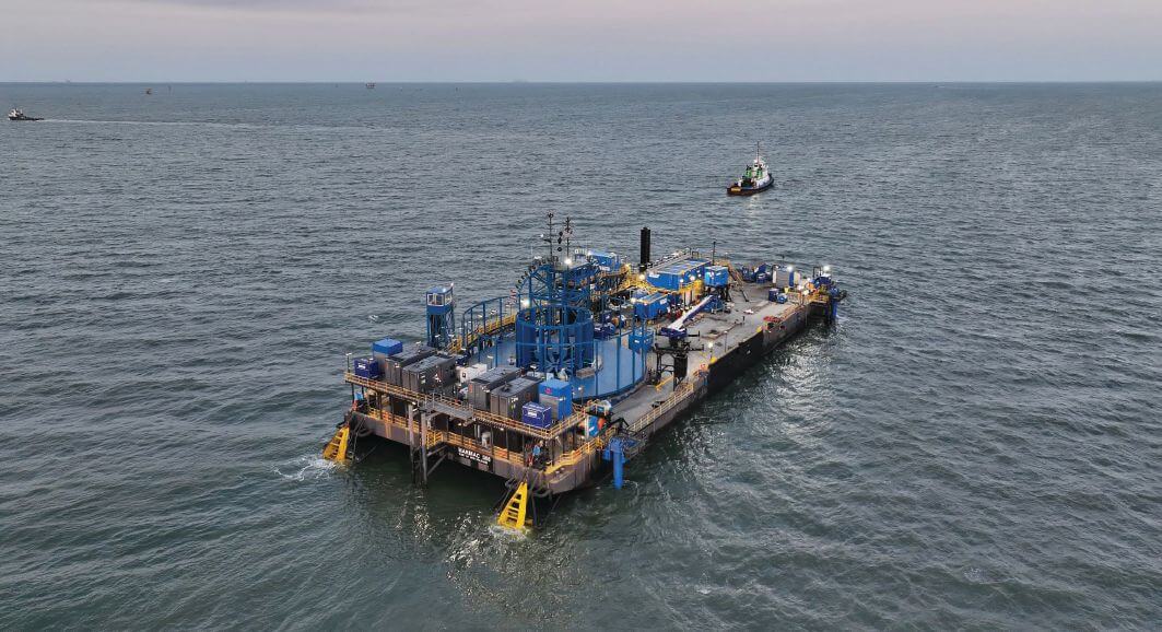 Aerial view of a cable lay barge at sea with a large blue cable carousel on deck and a small tug nearby.