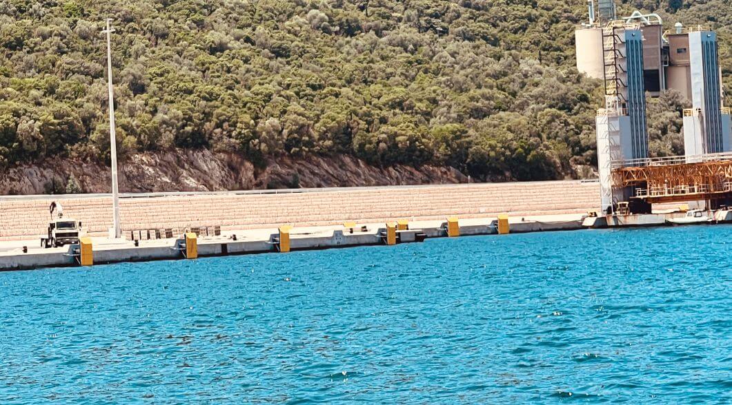 Long concrete quay at Igoumenitsa Port, Greece, fitted with evenly spaced yellow fender panels along the waterline below a wooded hillside.