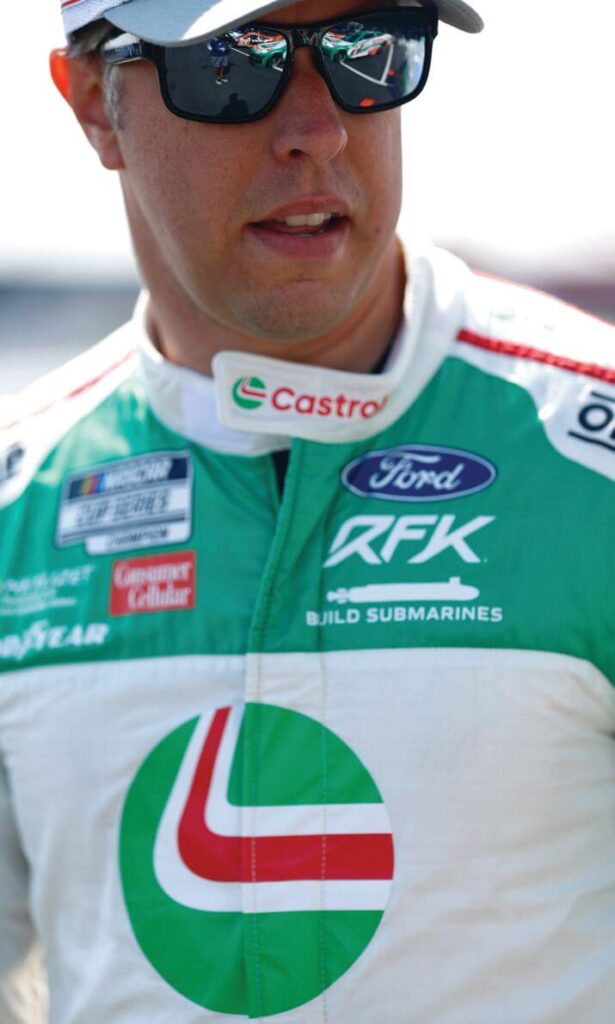 RFK Racing co-owner and driver Brad Keselowski in sunglasses and a green and white firesuit before a NASCAR Cup Series race.