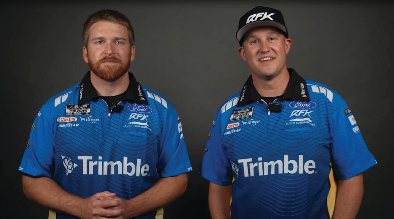 Two RFK Racing drivers, Chris Buescher (left) & Ryan Priest in blue Trimble team shirts standing in front of a dark backdrop.